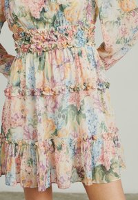 Blumenmusterkleid aus einem weichen, durchsichtigen Stoff in Pastellfarben. Es hat Rüschendetails an der Taille und am Saum sowie ein geschichtetes Design mit bunten Mustern.