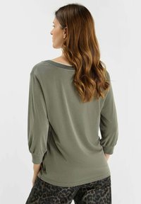Woman with long wavy brown hair wears a loose olive-green long-sleeve top and leopard-print pants, facing away against a white background.