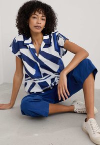 Camicia a maniche corte con colletto, decorata con un motivo astratto blu e bianco, abbinata a pantaloni cropped blu e sneakers beige, seduto su un pavimento in cemento.
