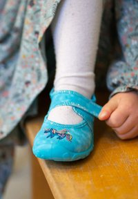 Ein türkisfarbener, stofflicher Mary Jane Schuh mit flacher Sohle zeigt eine Einhornstickerei und einen Klettverschlussriemen, getragen mit weißen Strumpfhosen.