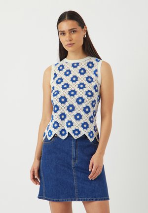 Vrouw draagt een mouwloze witte gehaakte top met blauwe bloemenpatronen en een blauwe denim rok, staand tegen een witte achtergrond.