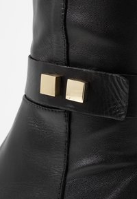 Bottine en cuir noir avec finition brillante, une large bride et deux accents carrés dorés. Des coutures détaillées sont visibles le long de la couture.