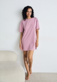 Rosa oversized T-skjortekjole med korte ermer, med et lite logo. Myk bomullstekstur og avslappet passform, som ender over kneet.