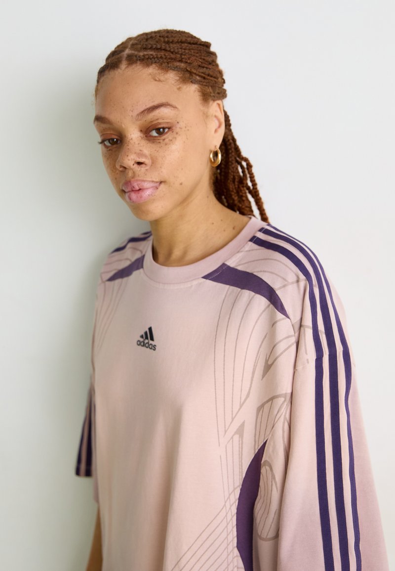 Jasnoróżowa, oversize koszulka z fioletowymi paskami, z abstrakcyjnym wzorem i czarnym logo Adidasa na piersi. Miękki, gładki materiał.