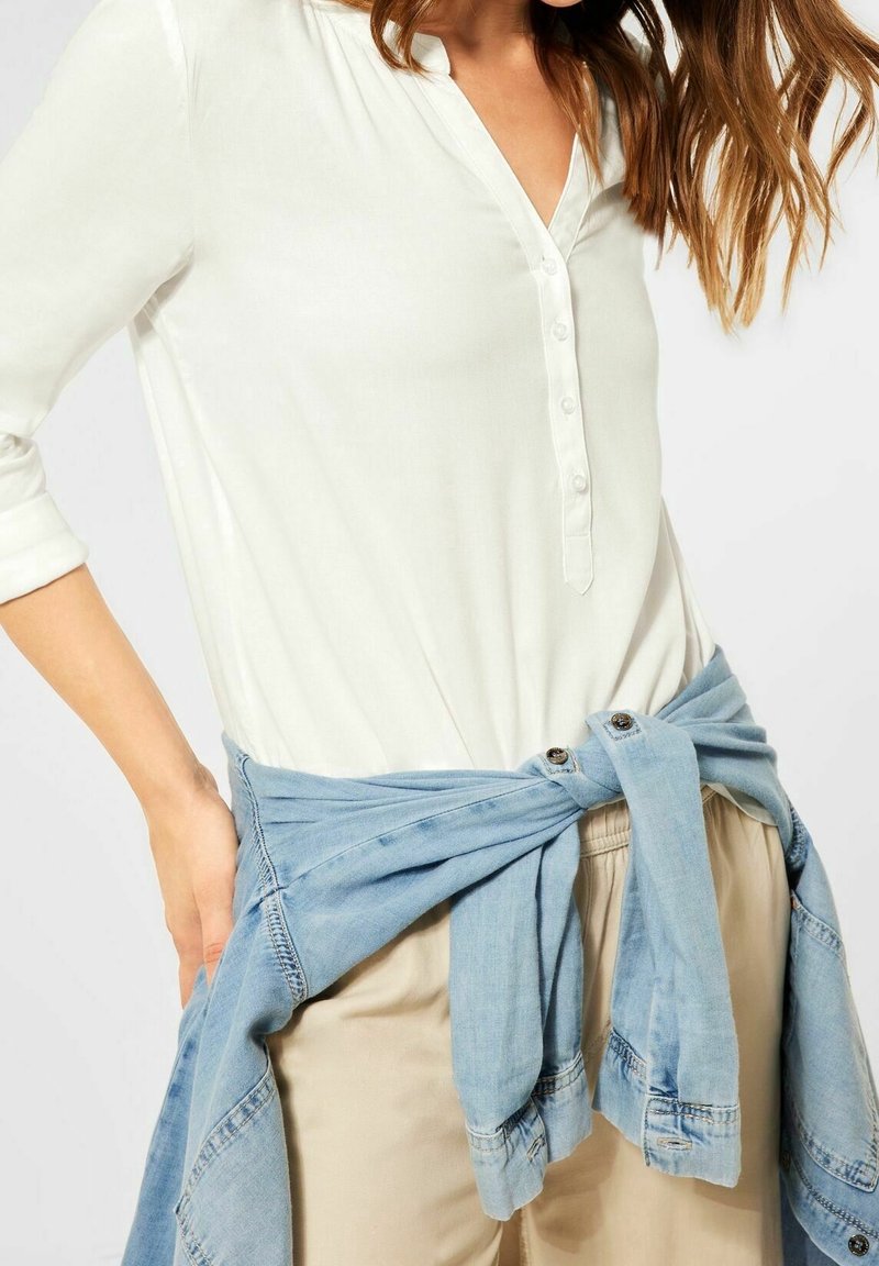 Camicia bianca con collo a V e maniche lunghe, abbinata a una camicia di jeans azzurra annodata in vita, sopra dei pantaloni beige.