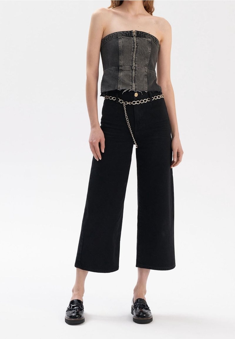 Femme portant un top en denim noir sans bretelles, un pantalon large noir avec une ceinture chaîne argentée et des mocassins noirs, debout sur un fond blanc.