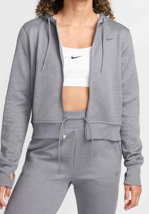 Woman wearing gray zip-up hoodie and matching joggers with drawstrings, paired with white sports bra featuring black logo.