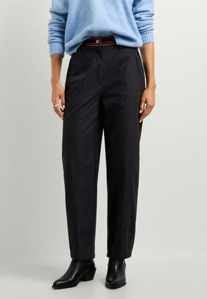 Personne portant un pantalon noir taille haute avec une ceinture rayée, un pull bleu clair et des bottines noires, debout devant un fond uni.