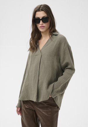 Žena oblečená v nadrozmernej ľanovej blúzke olivovo zelenej farby, tmavohnedých kožených nohaviciach a veľkých čiernych slnečných okuliaroch, stojaca s jednou rukou v vrecku.