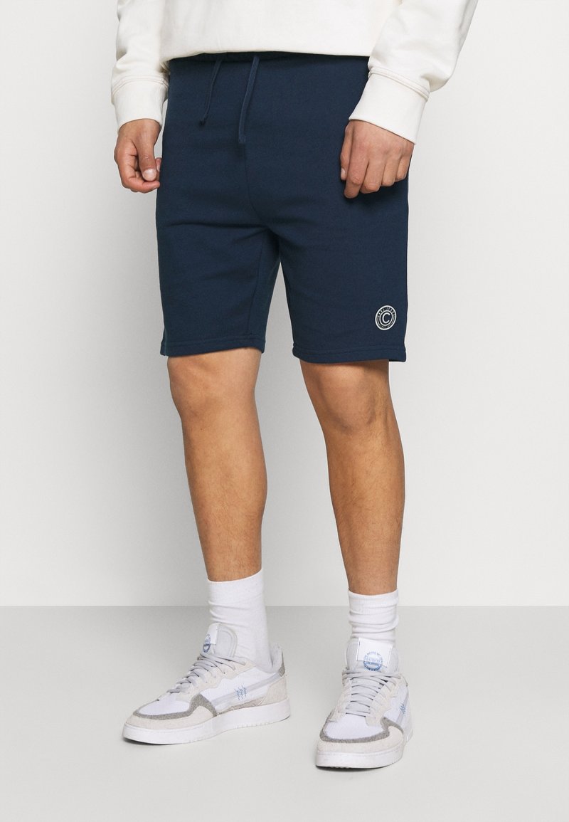 Granatowe szorty z mieszanki bawełny o luźnym kroju, z elastycznym pasem i białym logo na lewej udzie. Noszone z białymi trampkami.