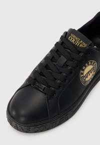 Melnas ādas tenisa kurpes ar teksturētām šņorēm, apaļu purngalu un gumijas zoli. Zelta "Versace Jeans Couture" logo uz sānu.