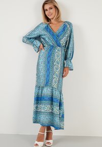 Pitkä sininen kuvioitu mekko, jossa on pitkät hihat ja V-pääntie. Sisältää kukka- ja paisley-kuvioita vihreänä ja valkoisena. Mekko ulottuu polvien alle.