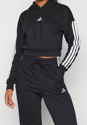 Czarna krótka bluza z kapturem z białym logo w kształcie trójkąta, z długimi rękawami z białymi paskami. W zestawie z czarnymi spodniami dresowymi w stylu jogger.