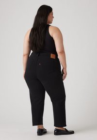 Jeans noirs taille haute avec une coupe droite, dotés de poches arrière et d'un patch en cuir. Portés avec un haut sans manches noir et des chaussures noires.