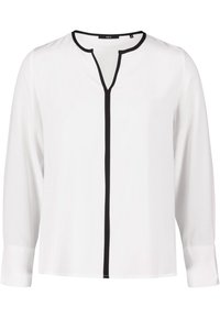 Blouse blanche à manches longues avec un décolleté en V et bord noir sur le devant. Tissu lisse sans motifs. Dispose de poignets avec boutons.