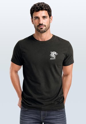Mann mit dunklem lockigem Haar, der ein schwarzes T-Shirt mit einem weißen brüllenden Leoparden und der Grafik "Nevertheless" auf der Brust trägt.