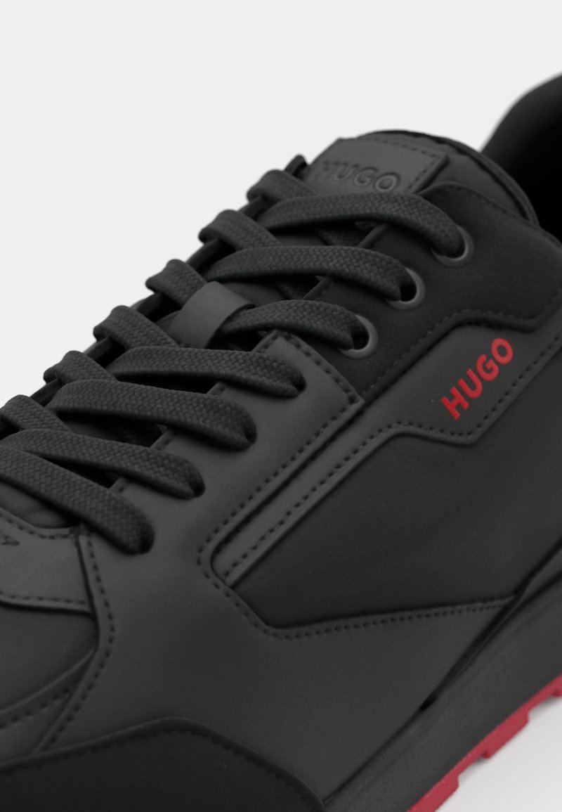 Scarpa sportiva nera con tomaia in pelle texturizzata, punta rotonda, lacci neri e accento del logo rosso. Presenta una suola ammortizzata con dettagli rossi.