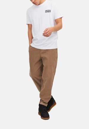 Weißes Baumwoll-T-Shirt mit schwarzem Logo, kombiniert mit braunen Cordhosen. Das Model trägt schwarze Schuhe mit Holzsohle.