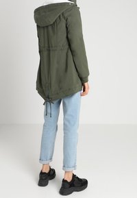 Parka verde oliva con capucha, cintura y dobladillo ajustables, y una textura suave. Lleva puesto sobre jeans de color azul claro y zapatillas negras.