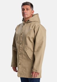 Un homme se tient de profil, portant un manteau imperméable beige avec une capuche, mettant en valeur un design simple et pratique sur un fond blanc uni.