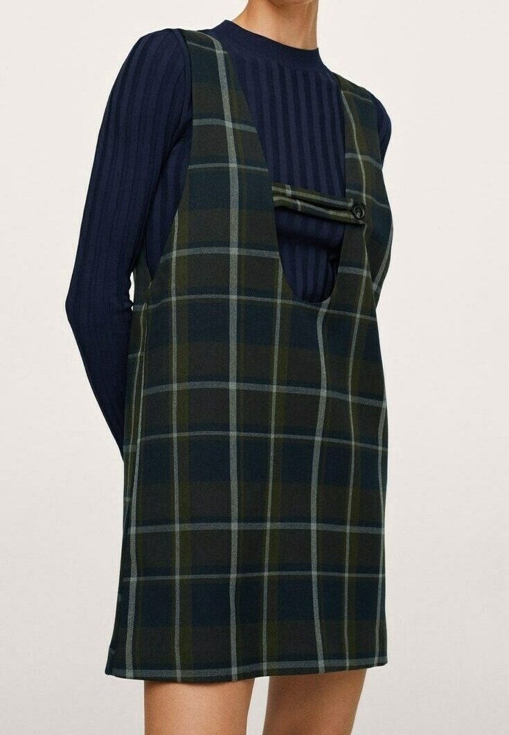 Femme portant une robe sans manches à carreaux vert foncé et bleu marine avec une découpe devant et un détail de boutons, par-dessus un haut à manches longues côtelé bleu marine.