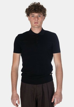 Giovane uomo con capelli ricci in piedi, indossa una polo nera a maniche corte e pantaloni plissettati marrone scuro su uno sfondo uniforme.