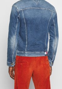 Džínová bunda středně modré barvy s prošisovanými místy, zapínáním na knoflíky a značkovou nášivkou na lemu. Kombinována s oranžovými manšestrovými kalhotami.