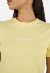 Tricou din bumbac galben deschis, cu guler rotund și un mic logo text brodat alb plasat în centru. Textură netedă.