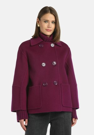 Cappotto doppiopetto in lana viola scuro, con grandi bottoni, due tasche frontali e un ampio colletto. Indossato sopra un dolcevita a coste.