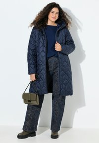 Abrigo largo acolchado de color navy con capucha, combinado con un suéter azul oscuro y pantalones con estampado. Lleva un bolso verde oliva; botas gruesas con suela negra.