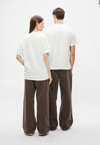 Fehér pamut rövid ujjú pólók, amelyeket két, háttal álló személy visel, lazán illeszkedő barna kordbársony nadrággal párosítva. Egyszerű kialakítás, minimális márkajelzés.