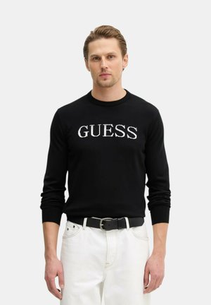 Mężczyzna ubrany w czarną koszulę z długim rękawem z logo "GUESS" oraz białe spodnie z czarnym paskiem, stojący na tle jednolitego jasnego tła.