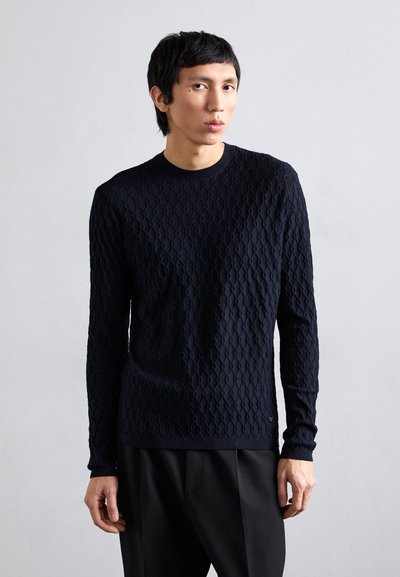 Pulover tricotat bleumarin, cu un model texturat în diamant, guler rotund și mâneci lungi. Asociat cu pantaloni negri croit.