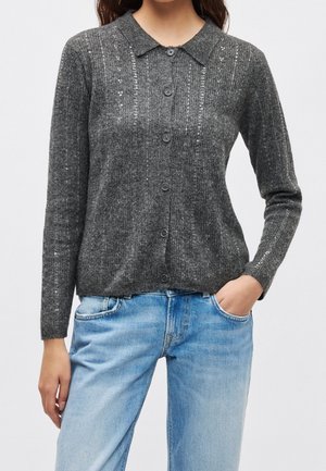 Femme portant un cardigan boutonné gris foncé avec de subtils détails en sequins et un jean bleu clair, une main dans la poche, debout devant un fond blanc.