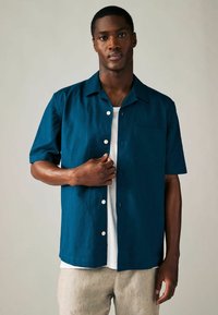 Kortärmad skjorta i mörk teal linne med en bröstficka, vita knappar och en avslappnad passform, bärs över en vit T-shirt.