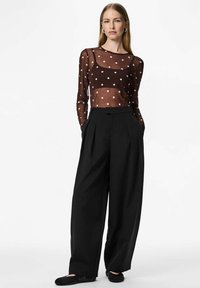 Maglia lunga trasparente marrone con pois bianchi, abbinata a pantaloni a gamba larga neri a vita alta e scarpe basse nere.