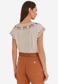 Haut gris clair à manches courtes avec un motif floral brodé marron sur l'épaule ; porté avec un pantalon taille haute couleur rouille doté d'une ceinture couture.