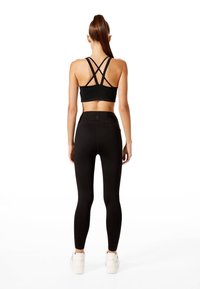 Soutien-gorge de sport noir avec des bretelles croisées, associé à un legging noir taille haute. Les deux présentent une texture lisse. Des baskets blanches complètent la tenue.