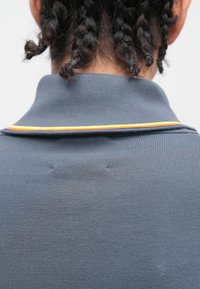 Camisola polo cinzenta com textura canelada e um detalhe decorativo colorido às riscas na gola. Vista de costas, mostrando tranças por cima.