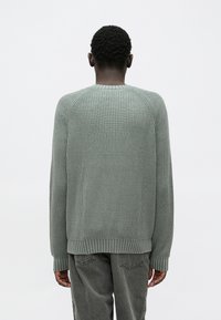 Zaļš ribots džemperis ar apaļu kaklu, noslīpētām plecu līnijām un raksturīgu materiālu. Dizainu papildina ribotas aproces un apakšmala.