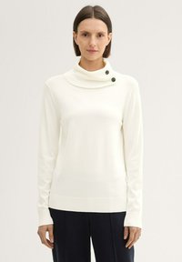 Pull blanc côtelé avec un col cheminée, comportant deux boutons noirs sur le col. Manches longues et ourlet droit. Porte avec un pantalon sombre.