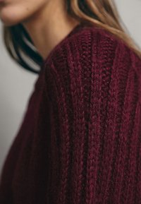 Burgundy neulepaita, jossa on pystyribbi kuvio, joka tuo esiin tekstuuria ja syvyyttä. Yksinkertainen pyöreä kaula-aukko ja pehmeä, paksu lanka.