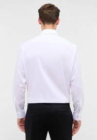 Camicia bianca a maniche lunghe con colletto classico, dotata di polsini con bottoni e una texture liscia e uniforme. Indossata con pantaloni neri.