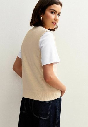 Mujer joven con cabello corto castaño, vistiendo un chaleco de punto sin mangas beige sobre una camisa blanca de manga corta y jeans oscuros, de perfil.