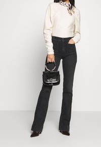 Schwarze, ausgestellte Jeans kombiniert mit einem cremefarbenen Langarmoberteil mit Puffärmeln, akzentuiert durch einen gepunkteten Schal und einer kleinen schwarzen Handtasche.