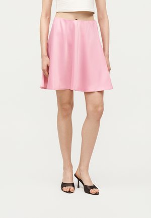 Γυναίκα που φοράει ροζ σατινέ φούστα σε σχήμα Α, λευκό κοντό τοπ και μαύρα ψηλοτάκουνα πέδιλα με ανοιχτά δάχτυλα, στέκεται μπροστά σε απλό φόντο.