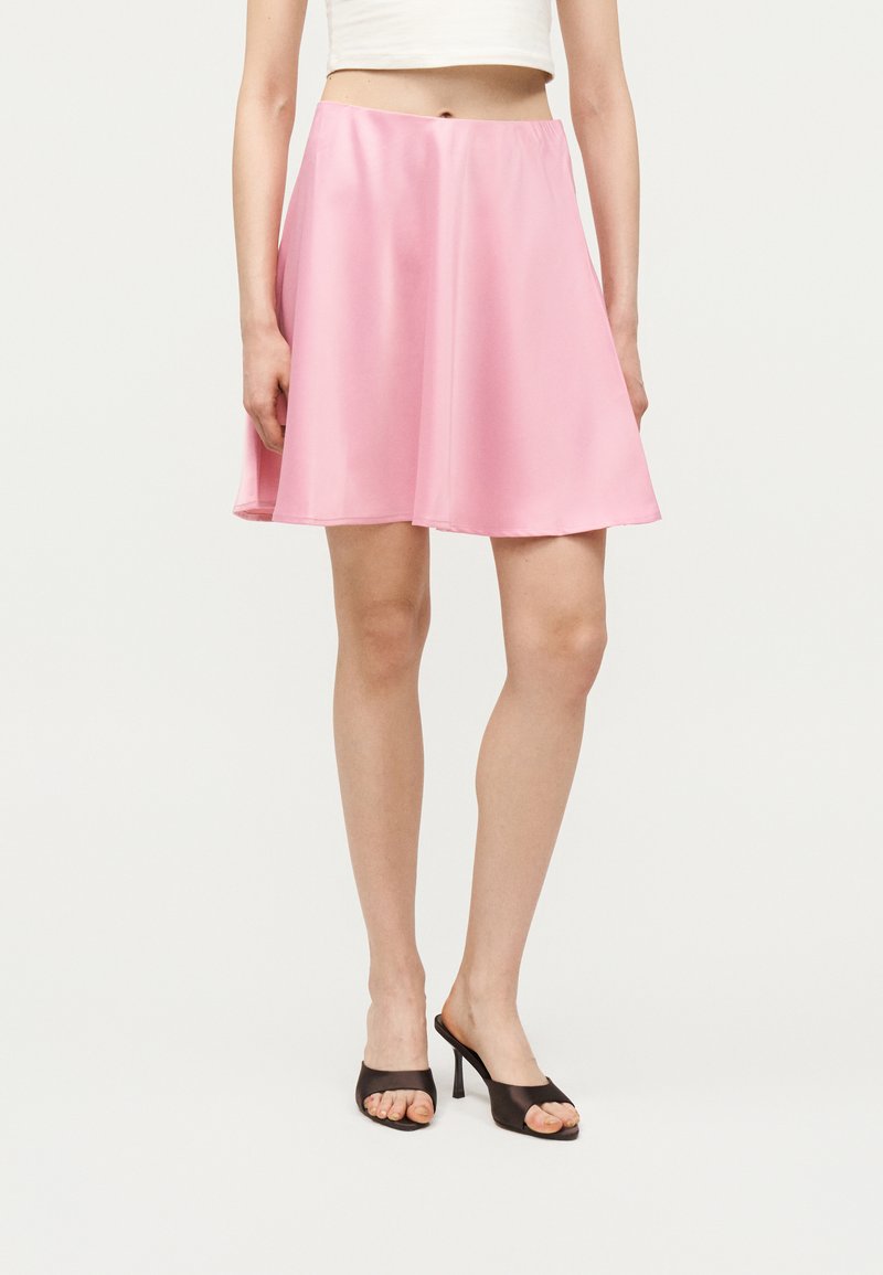 Γυναίκα που φοράει ροζ σατινέ φούστα σε σχήμα Α, λευκό κοντό τοπ και μαύρα ψηλοτάκουνα πέδιλα με ανοιχτά δάχτυλα, στέκεται μπροστά σε απλό φόντο.