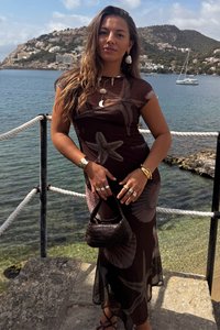 Vestido longo e transparente castanho com padrões de estrelas-do-mar, combinado com uma bolsa de mão tecida preta. Acessórios incluem brincos e vários anéis. Cenário do oceano.