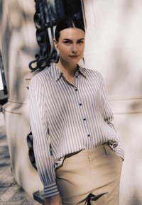 Sötét hajú nő csíkos gombos ingben és bézs nadrágban, az egyik kezét zsebre téve, világos kőfallal szemben áll a szabadban.
