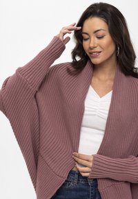 Strickjacke in staubrosa Farbe, mit langen Ärmeln, gerippter Textur, offenem Frontbereich und lässiger Passform. Über einem weißen Oberteil getragen.